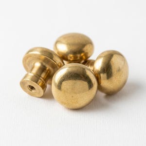 May include: Five brass cabinet knobs in a shiny gold finish. The knobs are spherical, with one having a cylindrical base and a threaded hole. The knobs are arranged on a white surface. Each knob is approximately 3 cm in diameter.