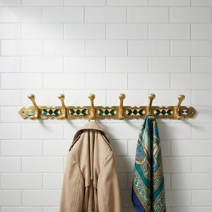 May include: A brass wall-mounted coat rack with seven hooks, featuring green and white geometric tile accents. A tan trench coat and a blue and green patterned scarf hang from the hooks, against a white tile wall.