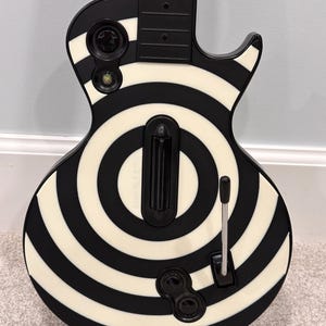 May include: A black and white electric guitar-shaped video game controller featuring a prominent bullseye pattern across the body. The controller includes a central black strum bar, a silver whammy bar, and circular control buttons on the face.