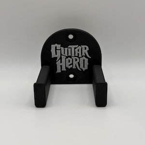 May include: Black guitar wall mount with the words "Guitar Hero" in gray lettering. The mount has two arms to hold a guitar and a semi-circular top with two holes for mounting.