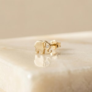 Könnte beinhalten: Ein goldfarbener Ohrstecker in Form eines Elefanten mit einem kleinen, klaren Edelstein als Auge. Der Ohrring ist an einem Stecker mit einem Butterfly-Verschluss befestigt. Der Elefant hat einen gebogenen Rüssel und große Ohren.