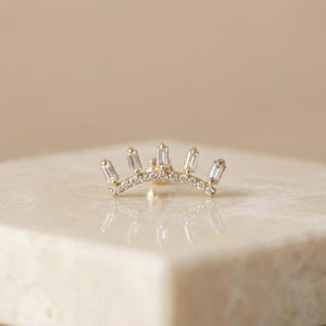 Könnte beinhalten: Ein goldfarbener Ohrschmuck mit geschwungenem Design. Er ist mit rechteckigen und runden, klaren Edelsteinen besetzt. Das Schmuckstück wird auf einer hellen Oberfläche präsentiert. Die Edelsteine sind klar und reflektieren das Licht.