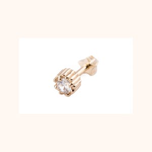 May include: A gold-colored stud earring with a clear, round gemstone. The earring has a decorative, ribbed setting around the gemstone and a post for insertion. The earring is set against a white background.