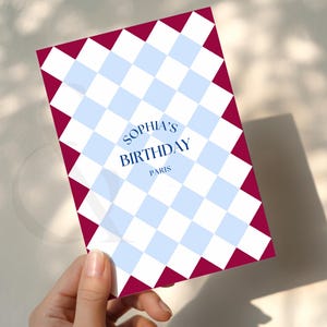 May include: A birthday card with a blue and white checkered pattern and a burgundy border. The card reads "Sophia's Birthday Paris" in a stylized font. The card is held by a hand.