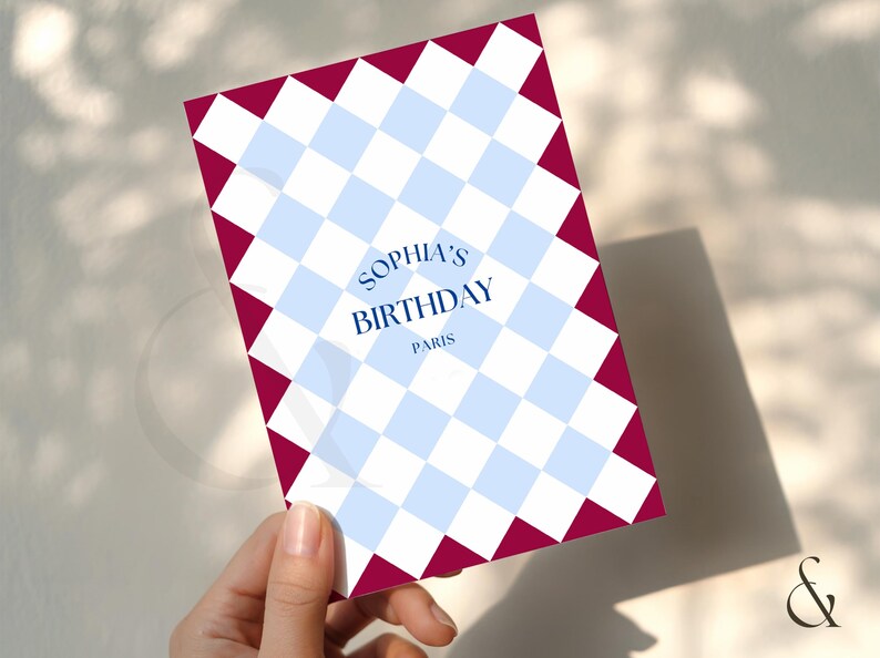 May include: A birthday card with a blue and white checkered pattern and a burgundy border. The card reads "Sophia's Birthday Paris" in a stylized font. The card is held by a hand.