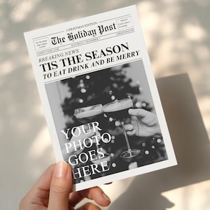 Puede incluir: Una impresión de periódico navideño en blanco y negro sostenida por una mano. El titular dice "TIS THE SEASON TO EAT DRINK AND BE MERRY". La imagen interior muestra dos manos brindando con copas de una bebida oscura. El texto en la impresión dice "YOUR PHOTO GOES HERE".