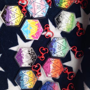 Può includere: Una collezione di portachiavi con dadi D20 in acrilico trasparente con chiusure rosse a forma di cuore. Ogni dado presenta una diversa combinazione di colori, tra cui arcobaleno, rosa, blu, giallo e viola, con i numeri da 1 a 20 visibili.