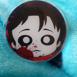May include: A round, clear button with a cartoon illustration of a person with dark hair and large eyes. The design includes a blood splatter on the face and a red border, set against a blue background.
