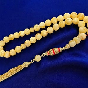 May include: A prayer bead necklace with creamy yellow beads. The beads are strung together with a silver-coloured chain and a decorative red and silver accent piece. The necklace ends with a gold tassel, set against a blue background.