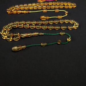 May include: Two amber-colored prayer beads, also known as a misbaha or tesbih, are displayed against a dark background. The beads are oval-shaped and strung together with yellow and green cords. The top strand has a yellow tassel.