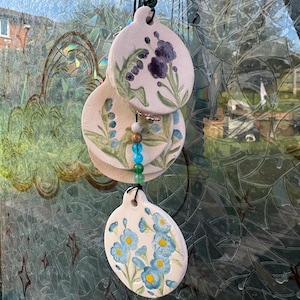 May include: A ceramic wind chime with oval pieces, each decorated with blue and purple floral patterns. The pieces are connected by a cord and beads, creating a decorative garden ornament. The measurements are in centimeters.