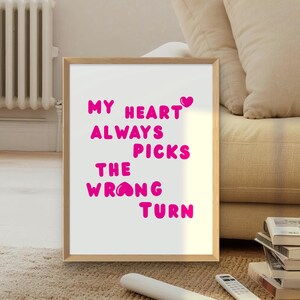 May include: A framed print with the text "MY HEART ALWAYS PICKS THE WRONG TURN" in bright pink letters. The artwork is in a light wood frame and is displayed on a rug in front of a couch.