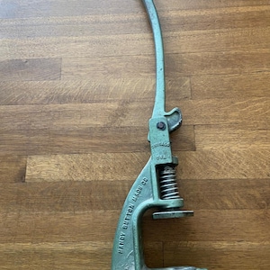 May include: A vintage green Handy Button Mach Co. button press. The press features a curved handle, a spring, and a base. The text "Handy Button Mach Co." is visible. The metal press shows signs of wear and tear, with a curved handle.