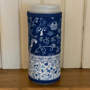 May include: A cylindrical, insulated water bottle sleeve with a white plastic lid. The sleeve is navy blue with a whimsical forest pattern in white. The bottom features a white and blue floral design.