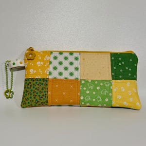 May include: A patchwork pencil case with a yellow zipper and a flower-shaped zipper pull. The case is made of squares of fabric in yellow, green, and orange with floral and geometric patterns. A green beaded charm hangs from the side.