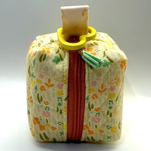 May include: A small, square, quilted pouch with a floral pattern in yellow, orange, pink, and green. It has a rust-colored zipper and a yellow loop with a small tassel. The pouch is designed for small items.