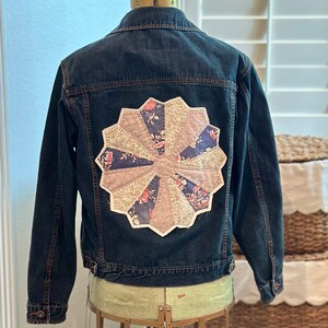 May include: A dark blue denim jacket with a large, circular patchwork design on the back. The patchwork is made of various fabrics in shades of pink, blue, and white, with floral patterns.