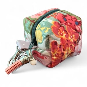 May include: A small, cube-shaped floral print pouch with a dark green zipper. The fabric features a mix of red, orange, pink, and green floral patterns. A small tassel and key ring are attached to the zipper.