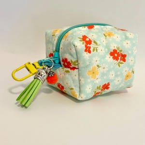 May include: A small, square, floral-patterned pouch with a teal zipper and a yellow clasp. The pouch features a light blue background with red, yellow, and white flowers. A green tassel and a small orange pom-pom dangle from the clasp.