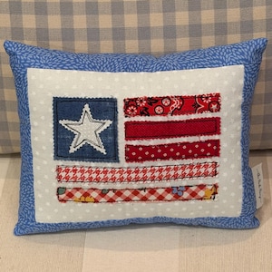 May include: A rectangular throw pillow featuring a white fabric center with a small star pattern and a blue fabric border. The center displays a flag-like applique design with a blue square containing a white star and five horizontal red fabric strips.