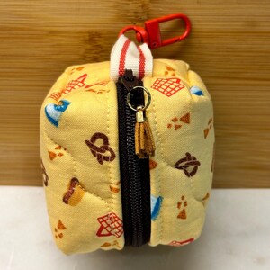 May include: A small, yellow, quilted pouch with a black zipper and a red clip. The pouch is decorated with pretzel, waffle, and other food-themed patterns. A small, brown tassel hangs from the zipper pull.