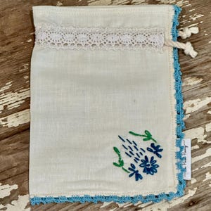 May include: Off-white linen pouch with a blue crocheted border and floral embroidery. The pouch features a white lace trim at the top and a drawstring closure. A small tag is attached to the side.