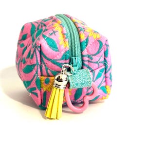 May include: A small, quilted pouch with a floral pattern in pink, teal, and yellow. The pouch has a teal zipper, a yellow tassel, and a pink heart-shaped clasp. The pouch is likely used for carrying small items.