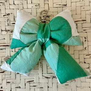 May include: A handmade bow-shaped keychain in shades of green and white. The bow is made of fabric patches and has a silver-colored metal clasp. The bow is a decorative accessory.