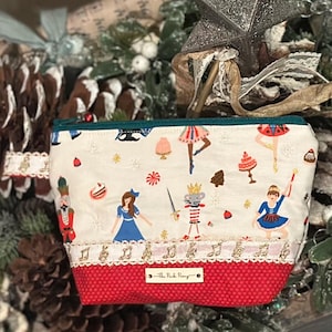 May include: A cream-colored cosmetic bag with a red bottom and a green zipper. The bag features a Nutcracker theme with illustrations of ballerinas, a mouse king, and musical notes. The text "The Pink Pony" is on the bottom.