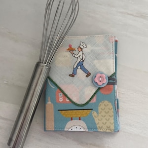 May include: A stainless steel wire whisk positioned next to a rectangular fabric cover featuring a light blue background with a repeating pattern of kitchen tools and an embroidered chef carrying a pie, secured by a pink flower-shaped button.