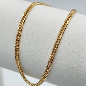 May include: A gold-colored curb chain necklace. The chain is displayed on a white surface. The links are closely interlocked, creating a smooth, continuous surface. The necklace has a polished finish, reflecting light.