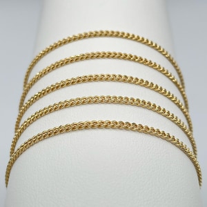May include: A collection of gold-coloured chain necklaces draped over a white form. The chains feature a curb link design, with a bright, shiny gold hue. The chains are arranged in parallel lines, creating a layered effect.