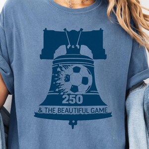 May include: A blue jean colored t-shirt featuring a dark blue graphic of the Liberty Bell with a soccer ball integrated into the design. The text 250 & THE BEAUTIFUL GAME is printed below the soccer ball graphic on the front of the shirt.