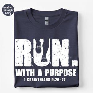 May include: A heather midnight navy t-shirt featuring a white distressed graphic that reads RUN. WITH A PURPOSE 1 CORINTHIANS 9:26-27. The letter U in RUN is replaced by a silhouette of legs and running shoes in a white, weathered, textured font.