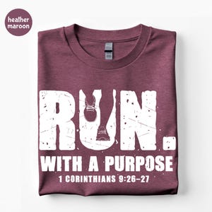 May include: A heather maroon short-sleeve t-shirt featuring white distressed text that reads RUN. WITH A PURPOSE 1 CORINTHIANS 9:26-27. The letter U in RUN incorporates a graphic of two running shoes. The shirt has a crew neckline and a tag.