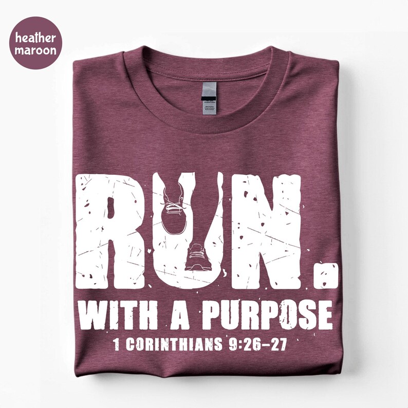 May include: A heather maroon short-sleeve t-shirt featuring white distressed text that reads RUN. WITH A PURPOSE 1 CORINTHIANS 9:26-27. The letter U in RUN incorporates a graphic of two running shoes. The shirt has a crew neckline and a tag.