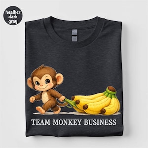 Puede incluir: Camiseta gris oscuro con un mono de dibujos animados tirando de un racimo de plátanos. El mono es marrón y sonríe. El texto "TEAM MONKEY BUSINESS" está impreso en blanco debajo de la imagen.
