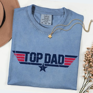 May include: A blue short-sleeve t-shirt with a distressed graphic print that reads TOP DAD in dark blue letters, flanked by red horizontal stripes and a dark blue star. The shirt features a Comfort Colors brand label at the inner neckline.