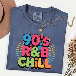 May include: A blue XL t-shirt with the text "90's R&B CHILL" in colorful bubble letters. A gold star pendant necklace rests on the shirt. The shirt is folded on a white surface.