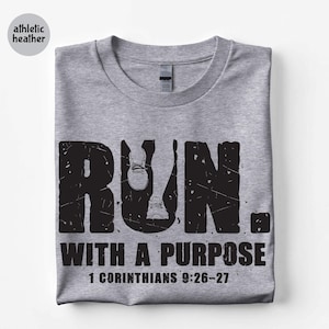 May include: An athletic heather grey t-shirt featuring a black distressed graphic that reads RUN with a shoe icon inside the letter U, followed by the text WITH A PURPOSE and the scripture reference 1 CORINTHIANS 9:26-27 in a bold, sans-serif font.