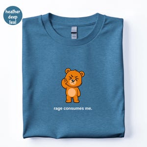 May include: A deep teal t-shirt with a cartoon bear illustration. The bear is orange with a grumpy expression and is making a hand gesture. The text "rage consumes me" is below the bear.