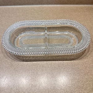 May include: Clear glass divided serving dish with an oval shape. The dish has a textured, beaded rim and a central divider. The interior features a dotted pattern. This vintage serving dish is suitable for serving snacks or appetizers.