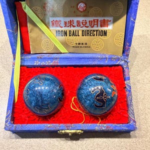 May include: A blue velvet box with a gold clasp contains two blue iron balls with gold designs. The box has a red interior and a gold-trimmed blue exterior. A small booklet inside reads "IRON BALL DIRECTION".