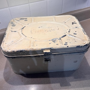 May include: A vintage, rectangular metal storage box with a cream-colored finish and distressed paint. The box has a hinged lid and a clasp closure. The top of the box has an oval design with the word "Sunbeam" embossed on it.