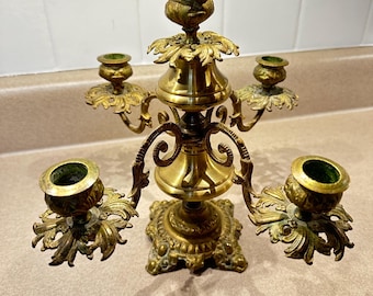 Puede incluir: Un candelabro de latón con cinco candeleros. El candelabro tiene una base cuadrada y detalles florales ornamentados. Los candeleros son circulares y tienen un interior verde. El diseño general es vintage.