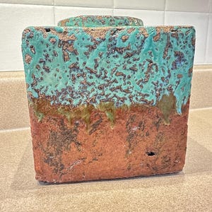 May include: A rectangular ceramic vase with a turquoise top and a reddish-brown bottom. The turquoise section has a textured, mottled appearance, while the bottom is a solid, earthy tone. The vase has a small, circular lid on top. The overall style is rustic and artistic.
