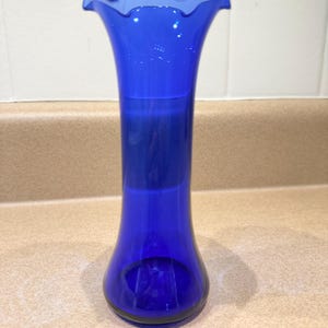 May include: A tall, cobalt blue glass vase with a ruffled rim. The vase has a slender, elongated shape, widening slightly at the base. The glass is smooth and reflects light, showcasing its vibrant color. A decorative piece for floral arrangements.