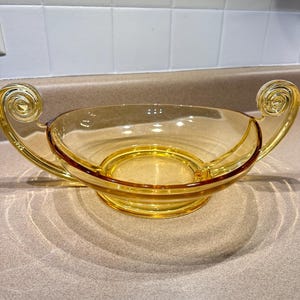 May include: A clear, amber-colored glass bowl with two decorative, swirled handles. The bowl has a circular base and a slightly flared rim. The glass has a smooth, glossy finish, and the bowl casts a shadow on the surface below.