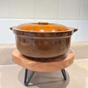 May include: A brown ceramic casserole dish with a lid, resting on a small wooden stand with black metal legs. The dish has a glossy finish and two side handles. The stand is round and made of light-colored wood.