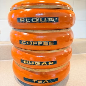 May include: An orange ceramic kitchen canister set with a lid and four stacked sections. Each section is labeled with black text: "FLOUR!", "COFFEE", "SUGAR", and "TEA". The canisters have gold-colored trim.